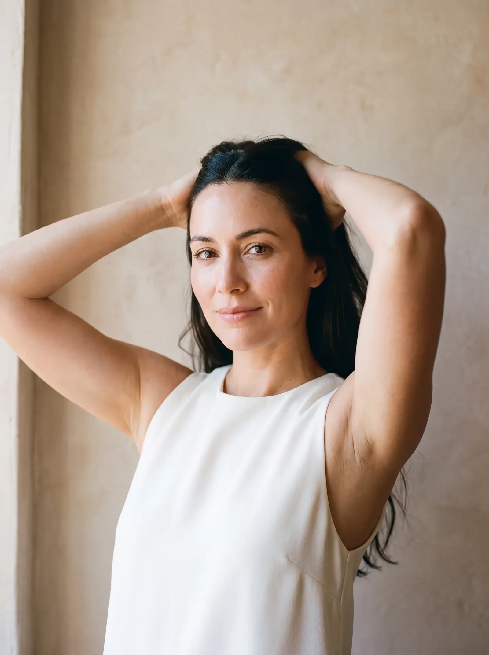 A woman in a white sleeveless top with her hands behind her head, in warm window light